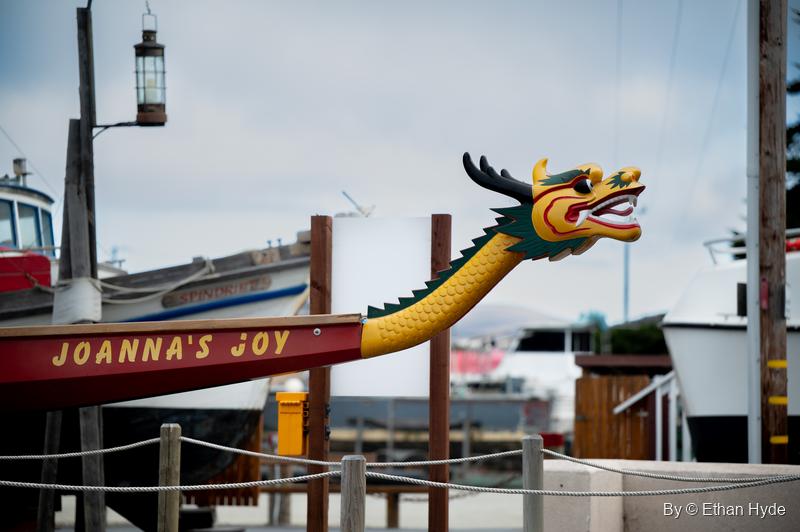 A dragon boat named Joanna's Joy with a carved yellow dragon prow, moored among working boats on the waterfront.