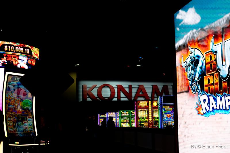 A red Konami sign glows in a dark booth between bright slot machine screens at G2E.