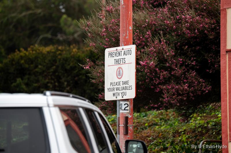 A red-and-white sign reading "Prevent auto thefts, please take your valuables with you" on a red post beside a parked SUV and flowering shrubs.