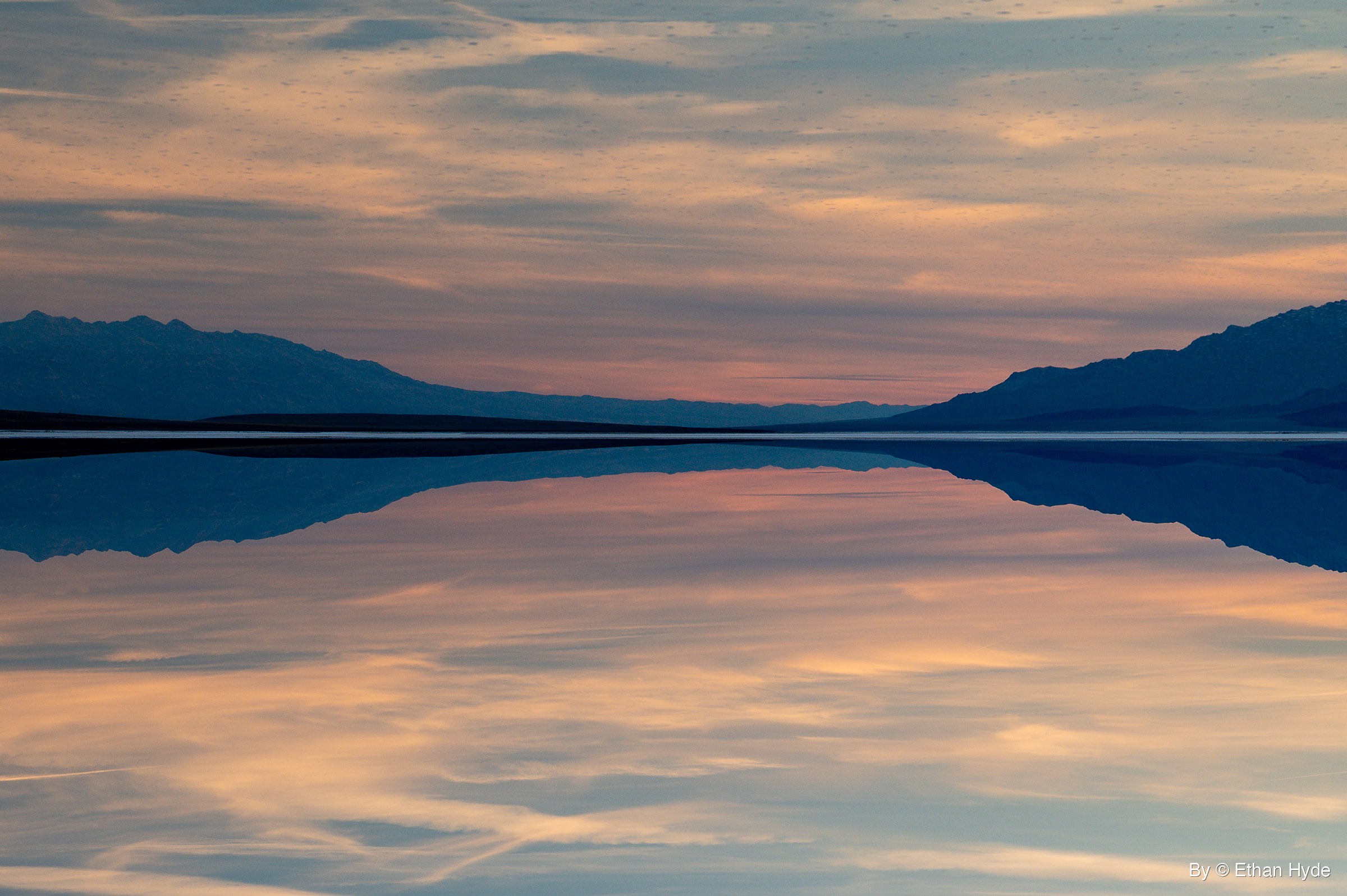 Calm standing water mirrors pink and gray sunset clouds and dark mountains, the frame shown upside down with the reflection on top.