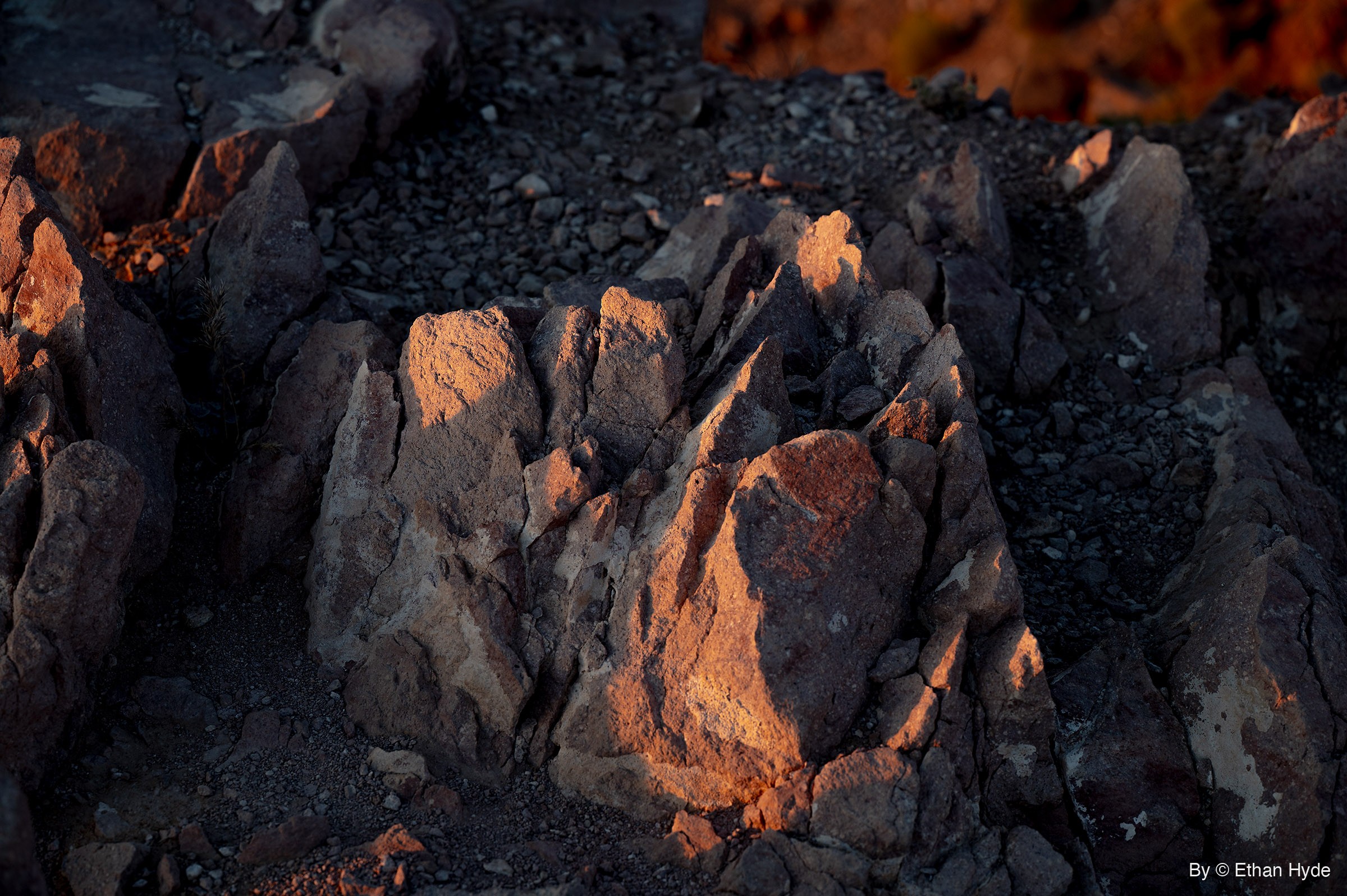 A rock outcrop broken into jagged upright blocks catches orange sunset light on a dark gravel slope, with a blurred orange hillside in the top corner.