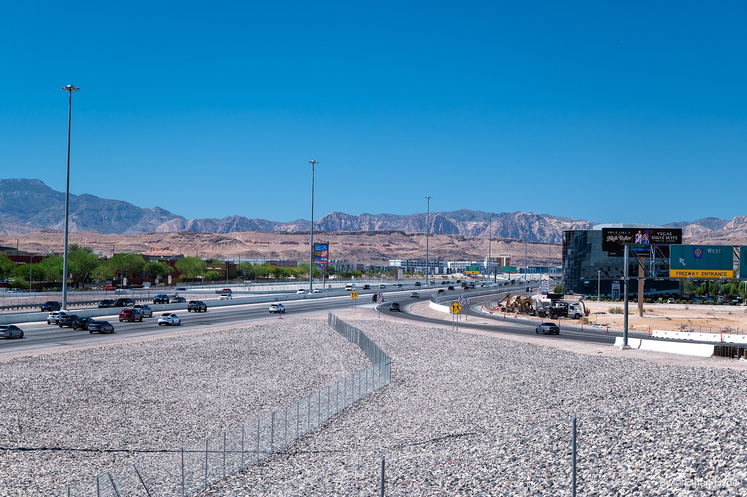 Wide view of a busy freeway and curving on-ramp beside a construction site, with a gravel slope and fence in front and rugged mountains beyond.