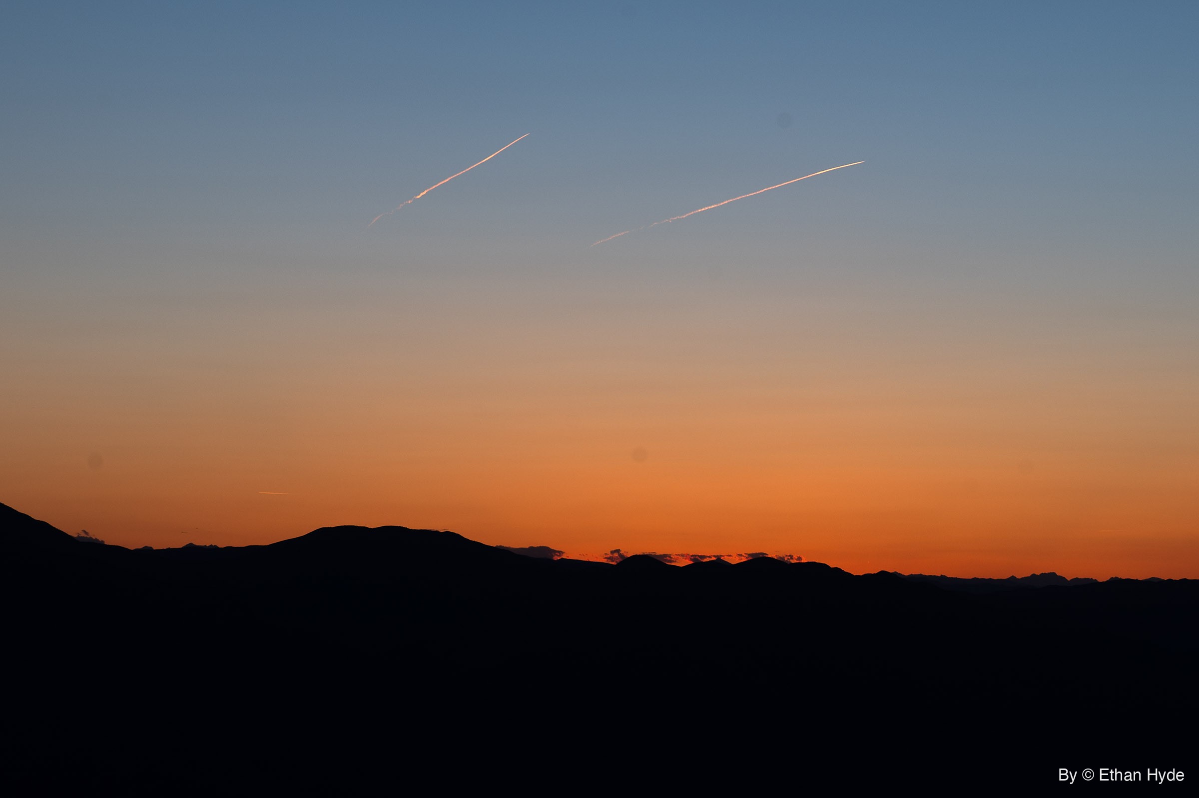 Two aircraft contrails cross a blue-to-orange twilight sky above a black silhouetted mountain ridge.