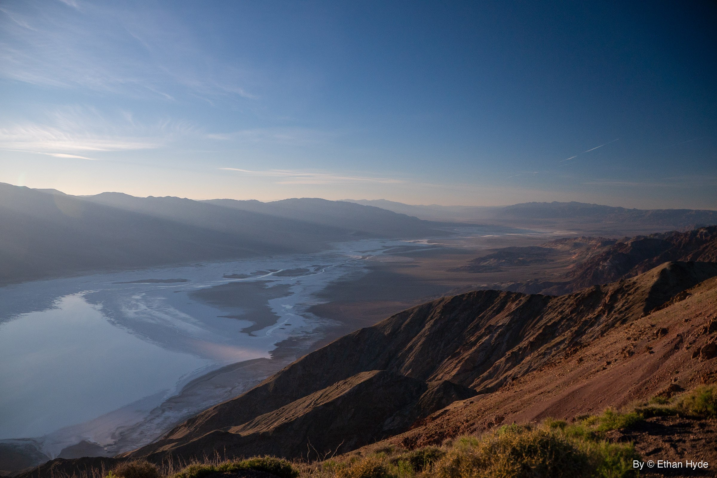 Looking north from a high brushy ridge down Death Valley, with shallow water spread across the salt flat and hazy ranges beyond.