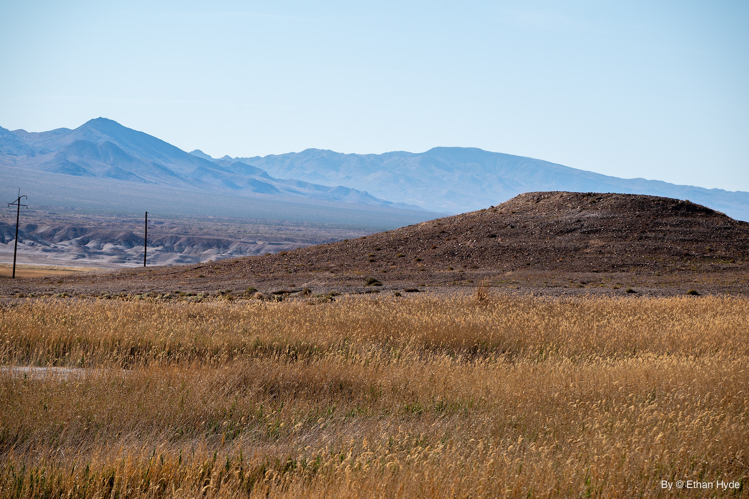 A field of dry golden grass in front of a brown dirt mound, with pale eroded badlands, two power poles and hazy blue mountains behind.