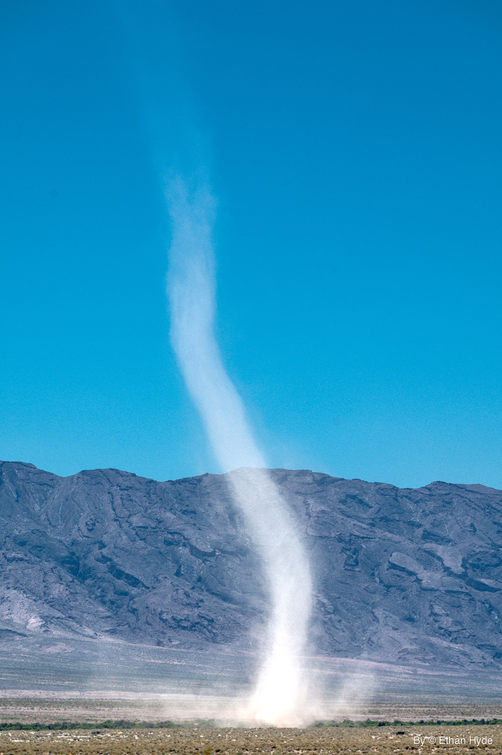 A tall pale dust devil rises from a desert floor, bending as it climbs past a gray mountain ridge into clear blue sky.