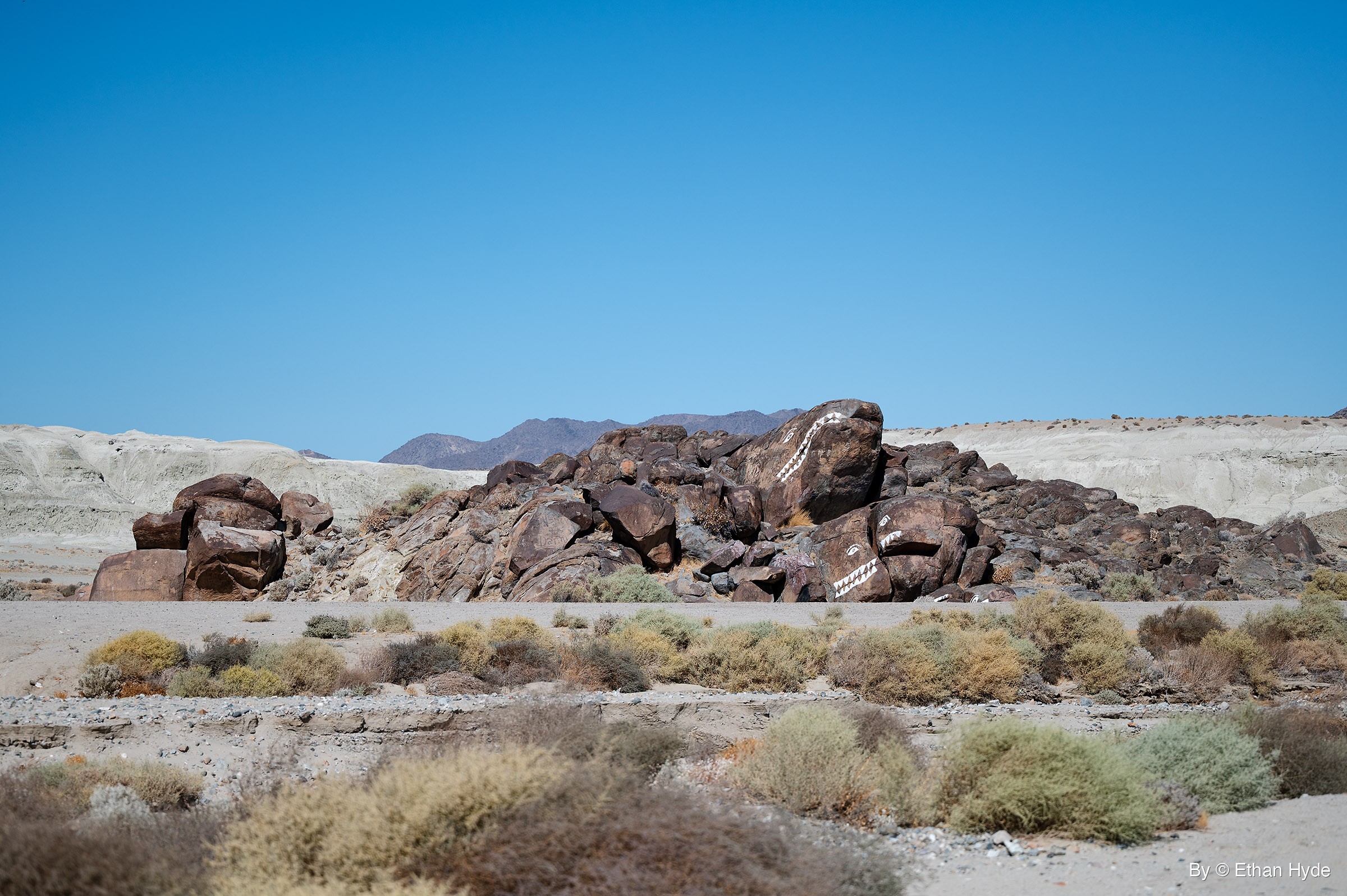 A pile of dark boulders painted white to look like fish sits beside a dirt road in a pale desert wash, with low mountains behind.