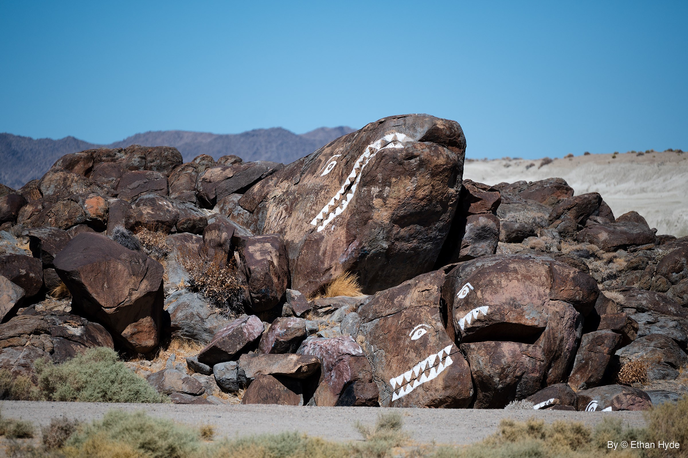 Dark boulders painted with white eyes and zigzag teeth so they look like fish, piled beside a gravel road under a clear blue sky.