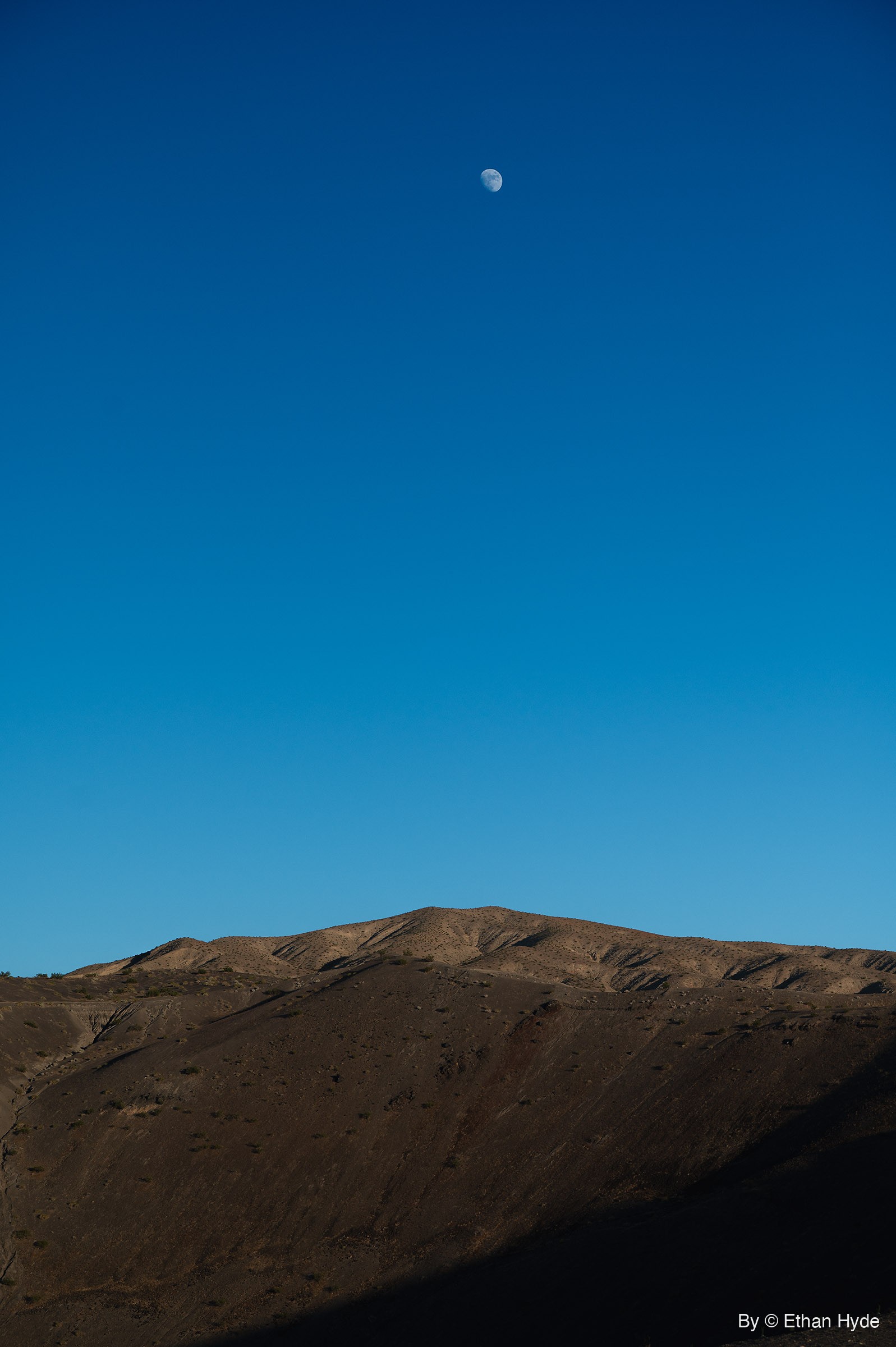 A small moon hangs high in a deep blue sky above a bare desert ridge whose near slope lies in shadow.