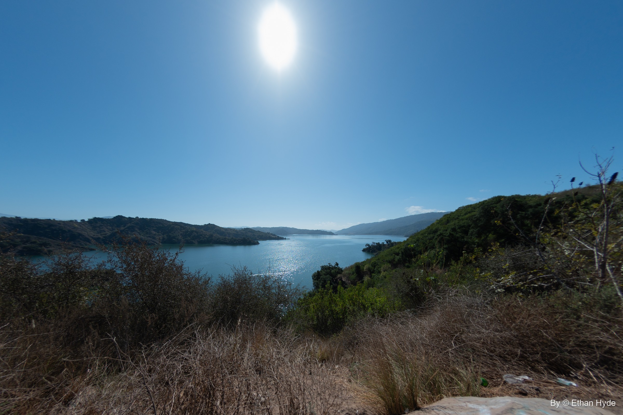 A fisheye view down a dry, scrubby slope to the blue arms of Lake Casitas, with the sun blazing overhead.