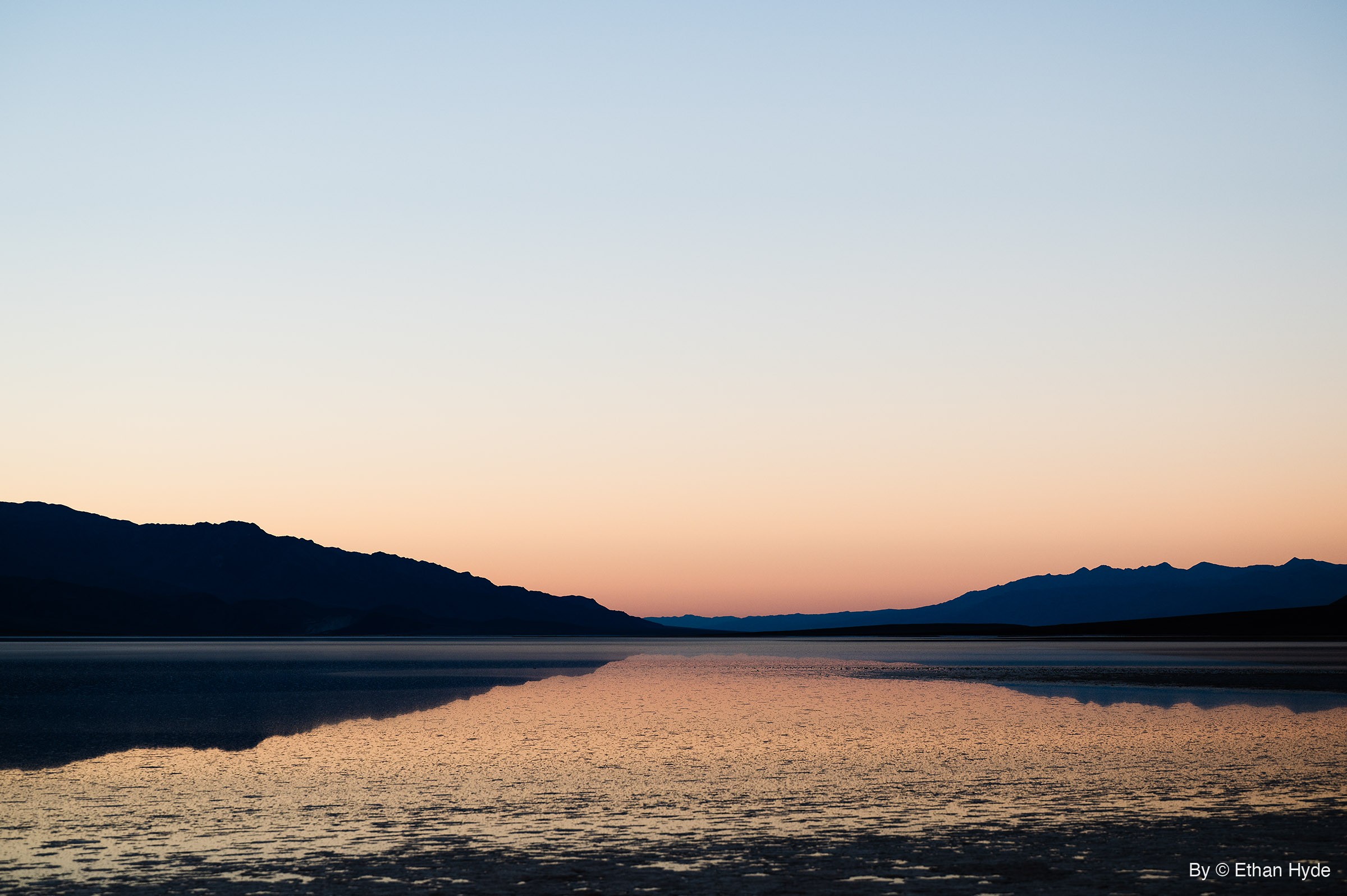 Shallow floodwater over the salt flat reflects an orange and blue sunset sky between two dark mountain ridges.