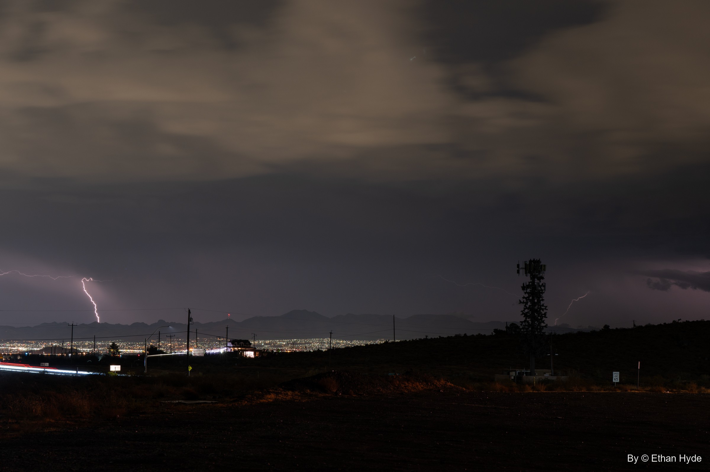 A lightning bolt strikes beyond a spread of city lights at night, with a cell tower silhouetted on the dark ridge.