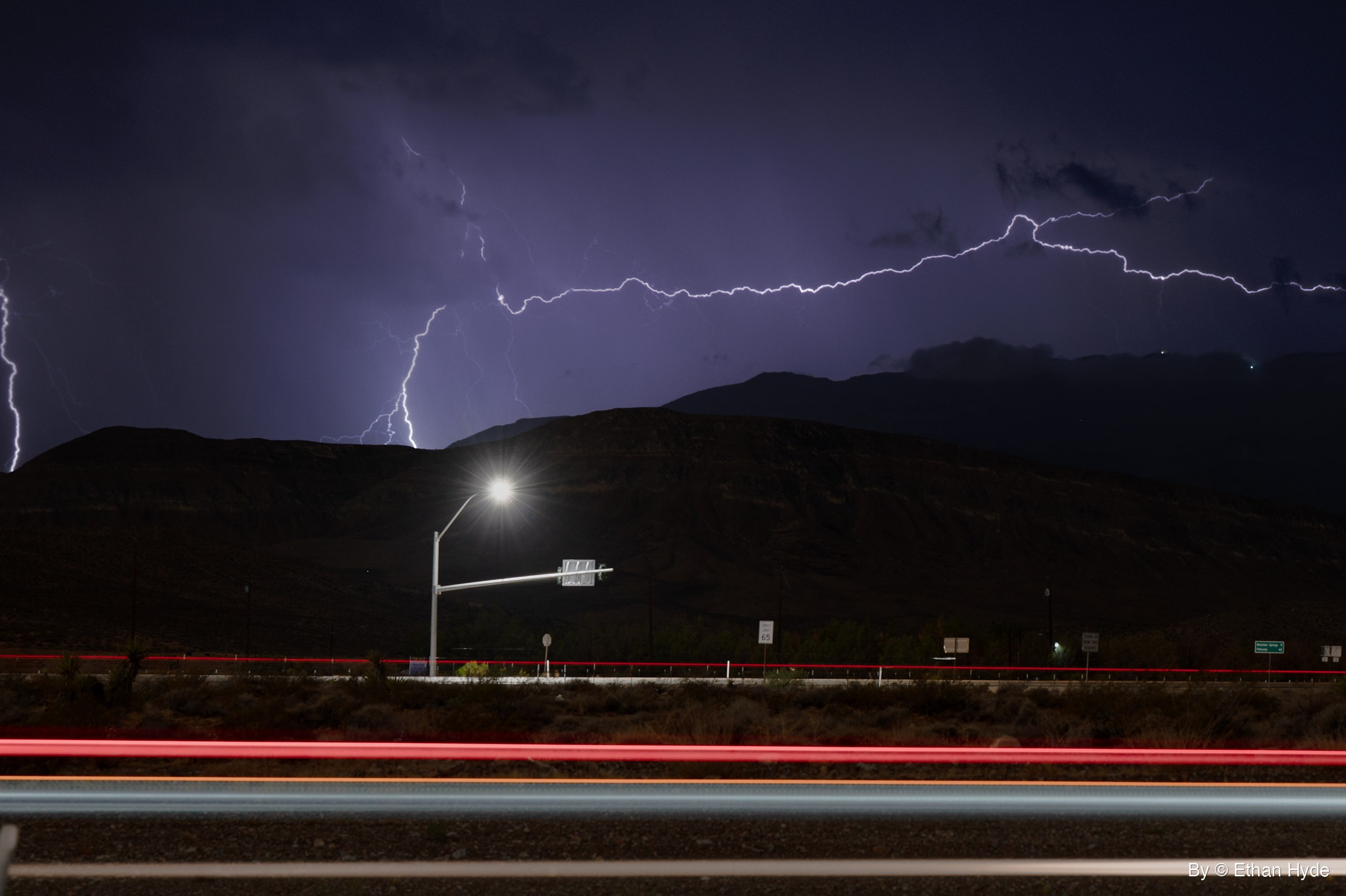Lightning branches across a purple night sky above dark mountains, with a lit streetlight and red and white car light trails along a road below.