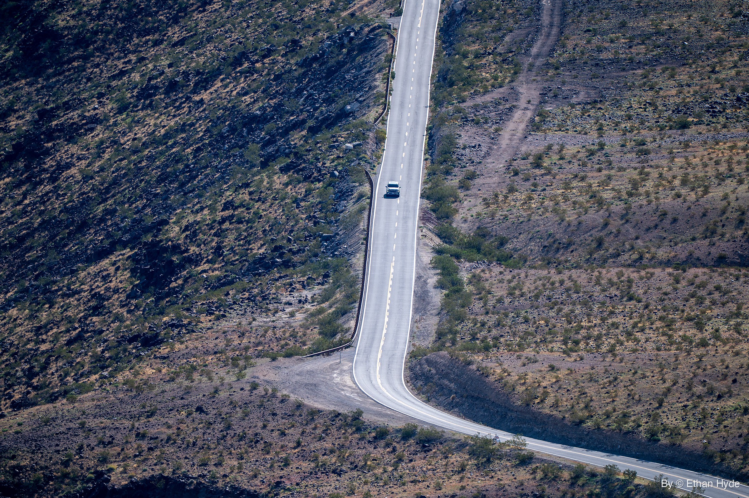 A single white car travels a two-lane road running straight up a scrubby desert slope, with a guardrail on the left and a curve at the bottom.