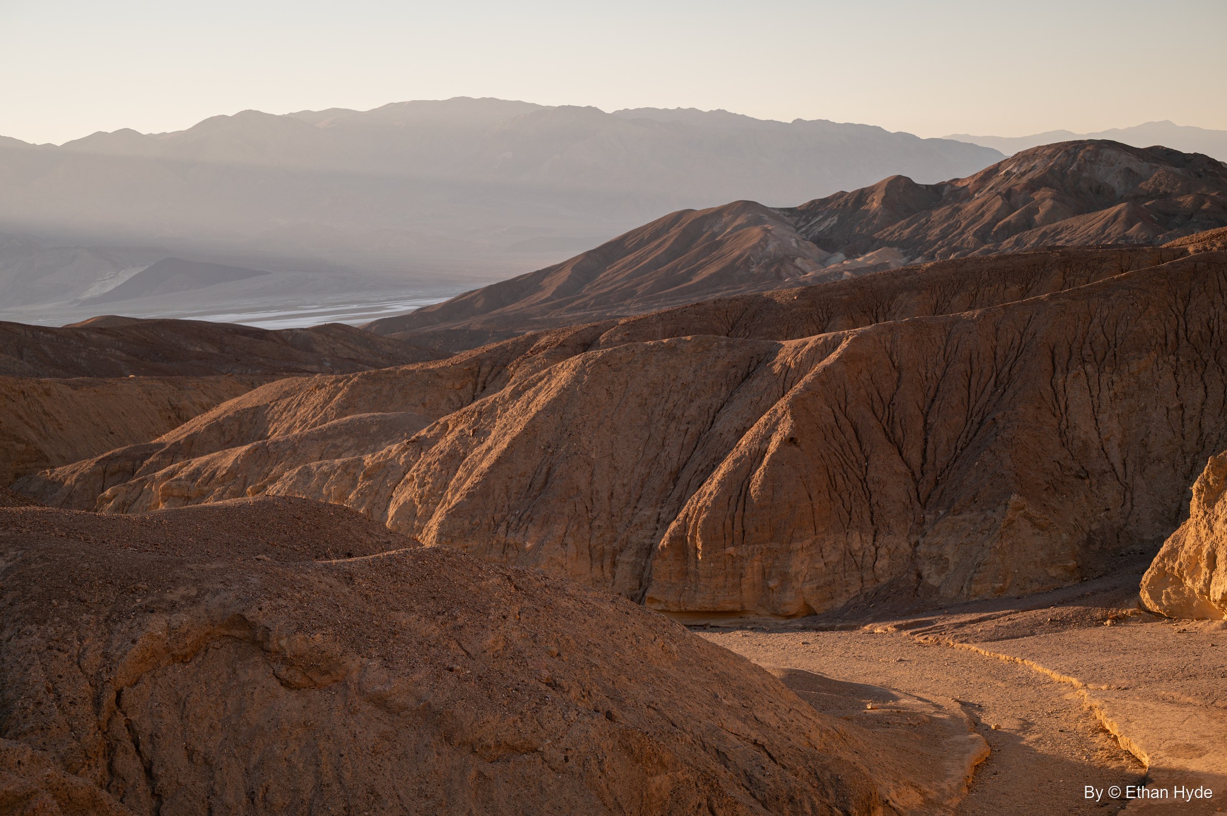 Low sun lights brown, eroded hills off Artists Drive, with the valley floor and a far mountain range in haze beyond.