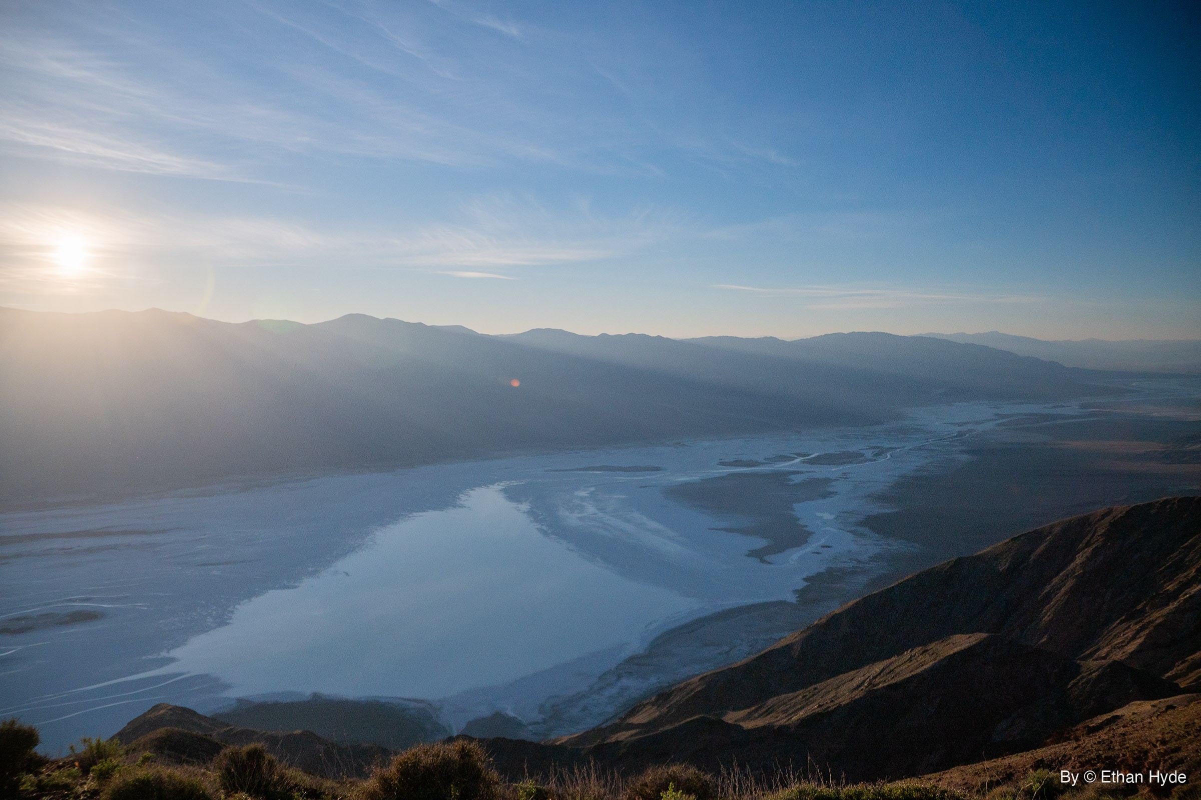 The sun hangs low over a hazy mountain range above Death Valley's salt flat, which is covered in shallow water, seen from a brushy ridge.