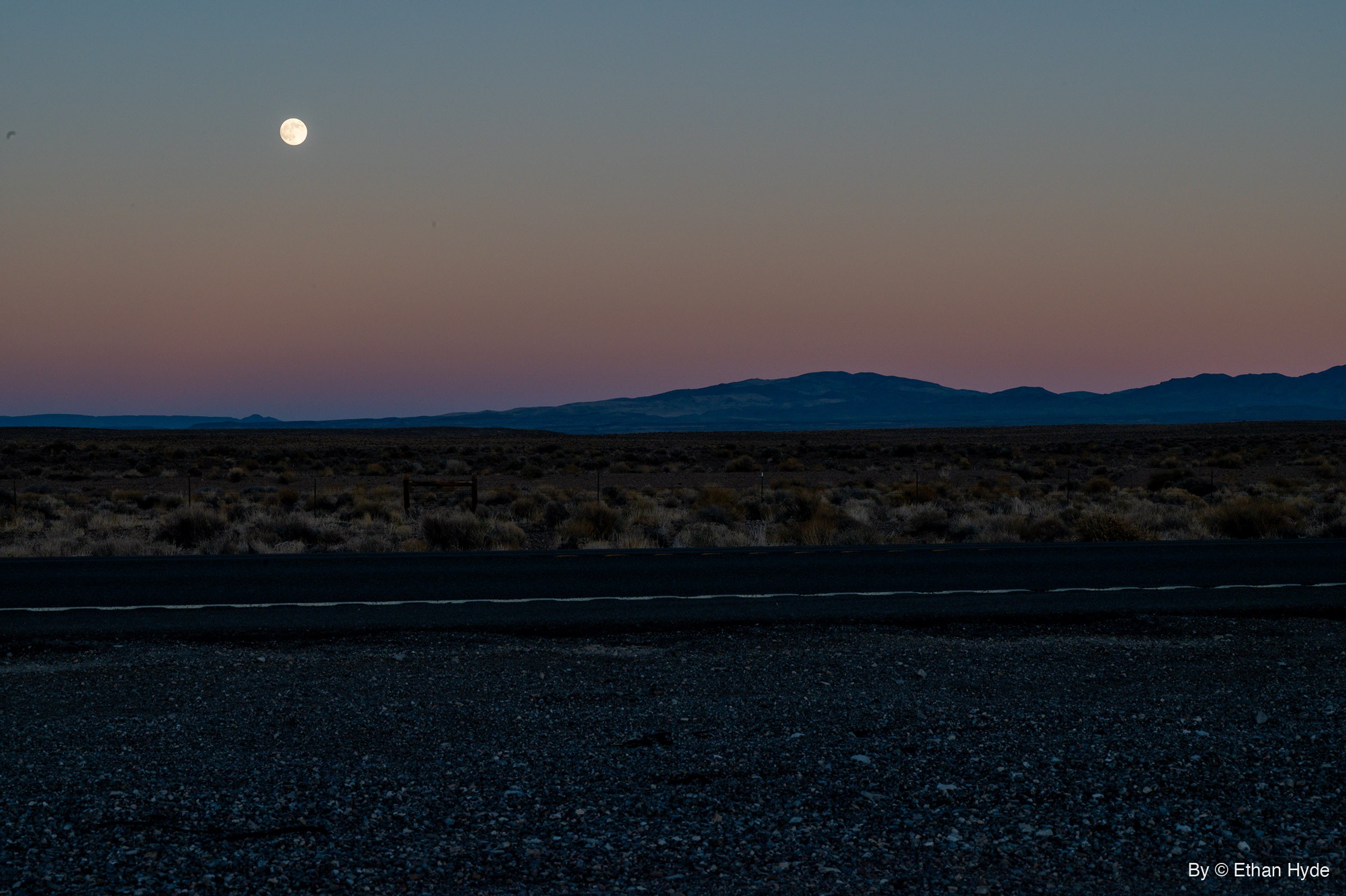 A near-full moon rises into a pink and blue dusk sky above a low mountain range, sagebrush flats and a paved highway with a gravel shoulder.