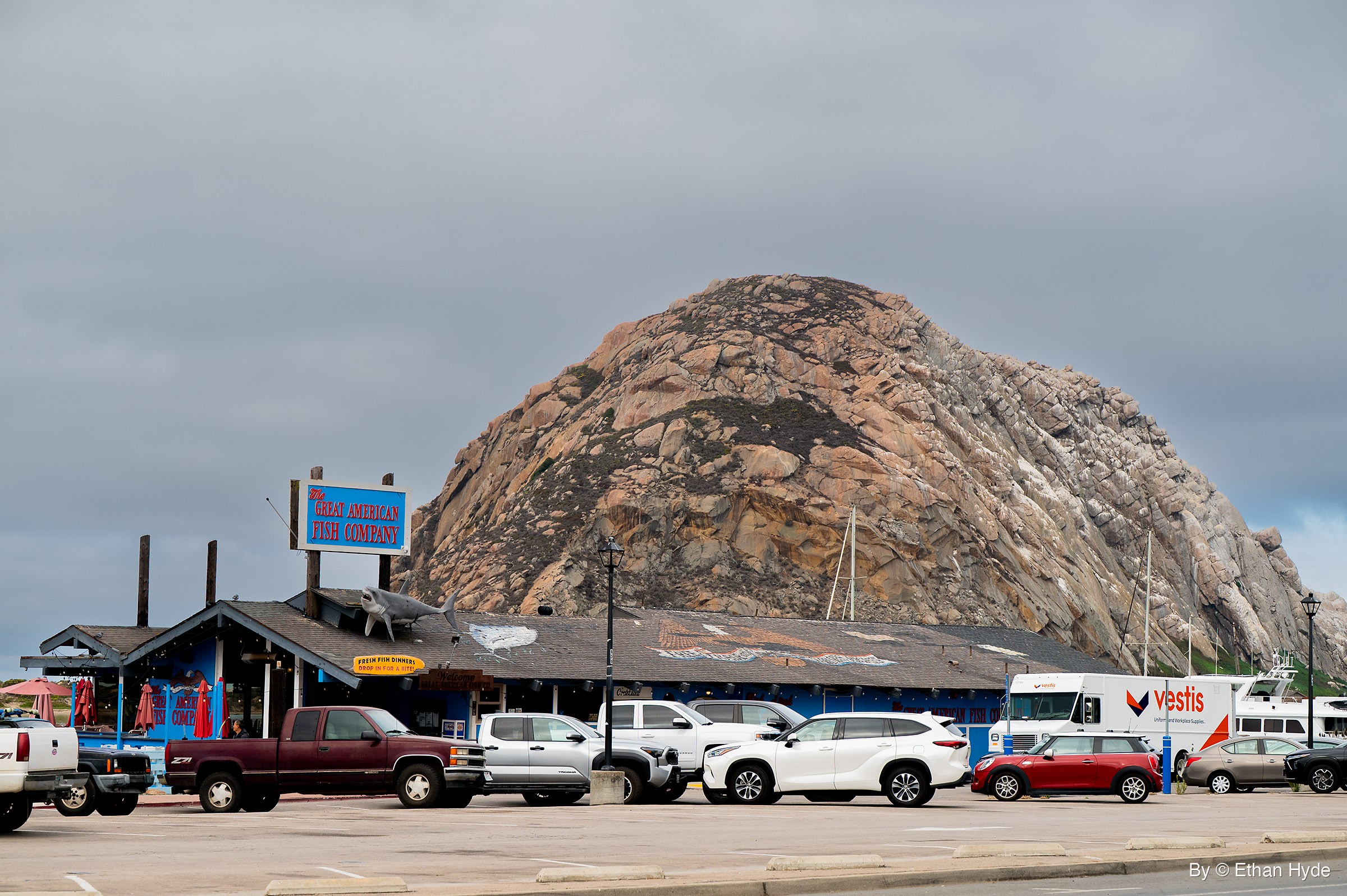 Morro Rock's rounded dome rises behind a blue waterfront seafood restaurant and a parking lot of cars and trucks under an overcast sky.