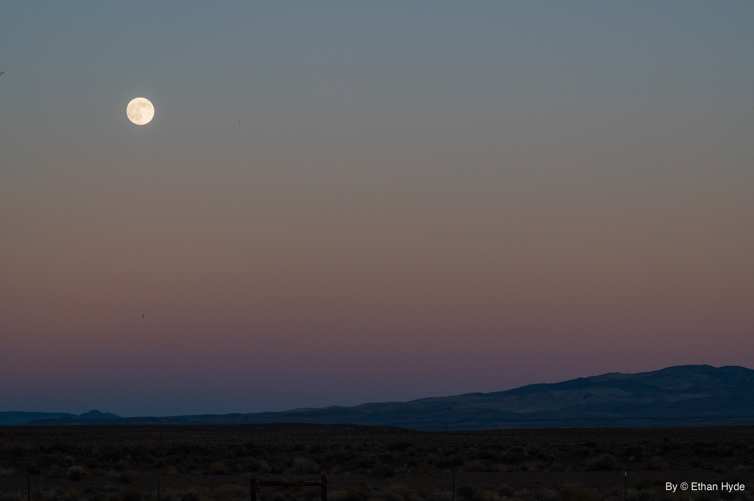 A nearly full moon high in a blue-to-pink twilight sky above a dark mountain ridge, a brushy flat and a fence line.