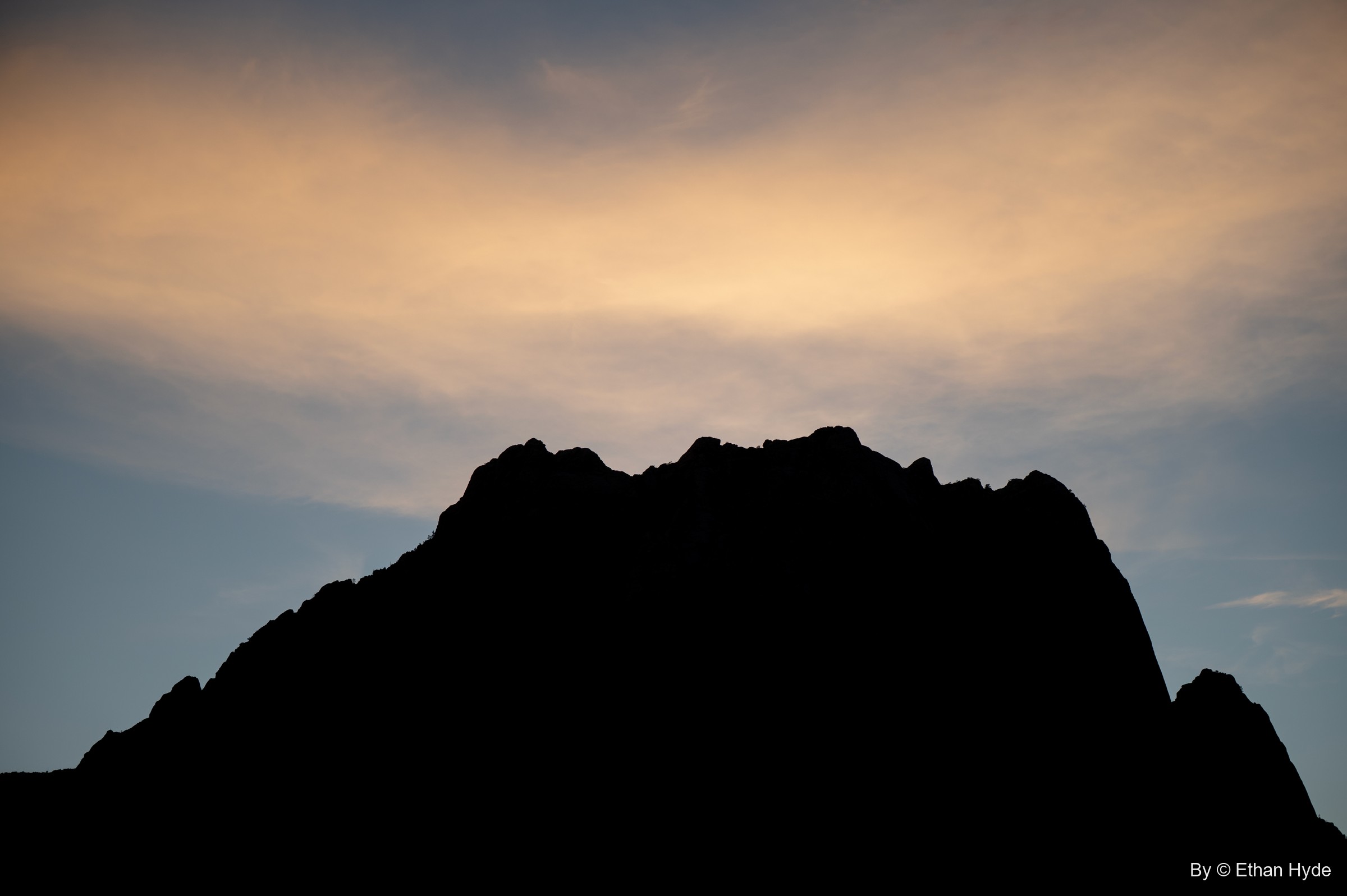 A jagged mountain peak in black silhouette under a warm, cloudy sunset sky.