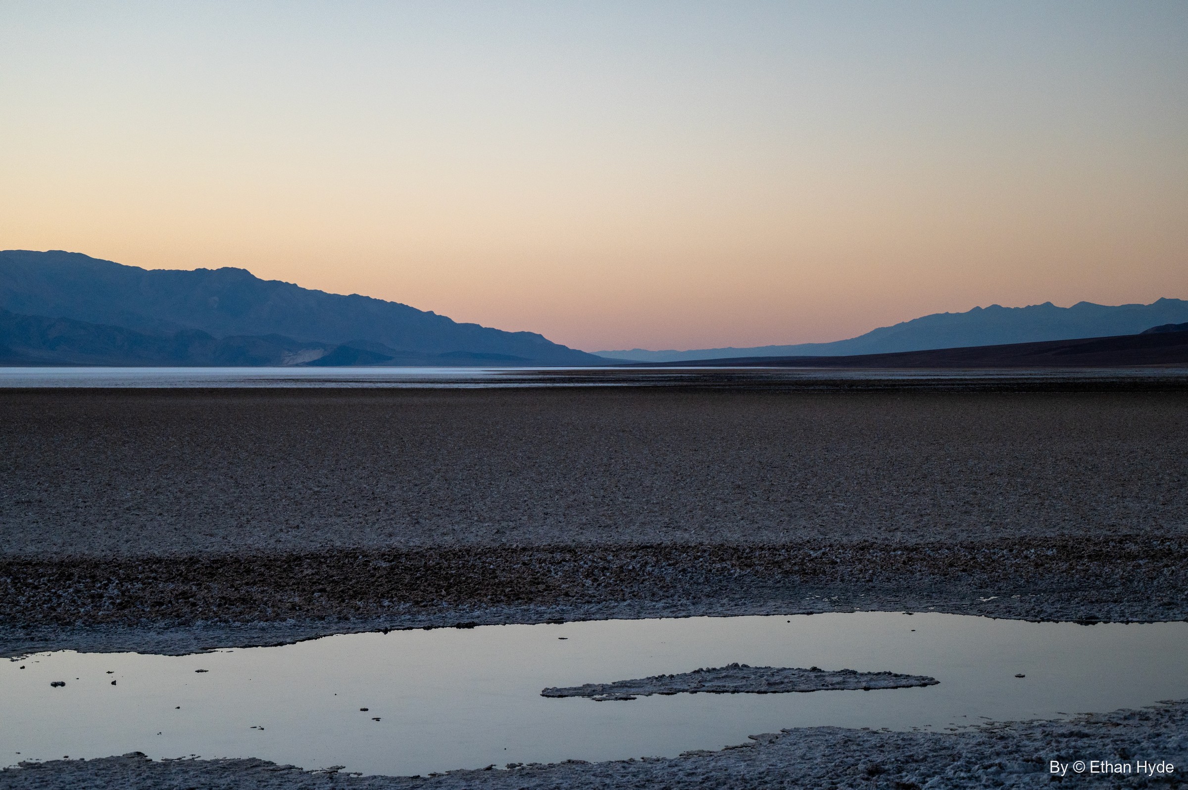 A shallow pool on the Badwater salt flat holds the pink and blue dusk sky beneath distant mountains.