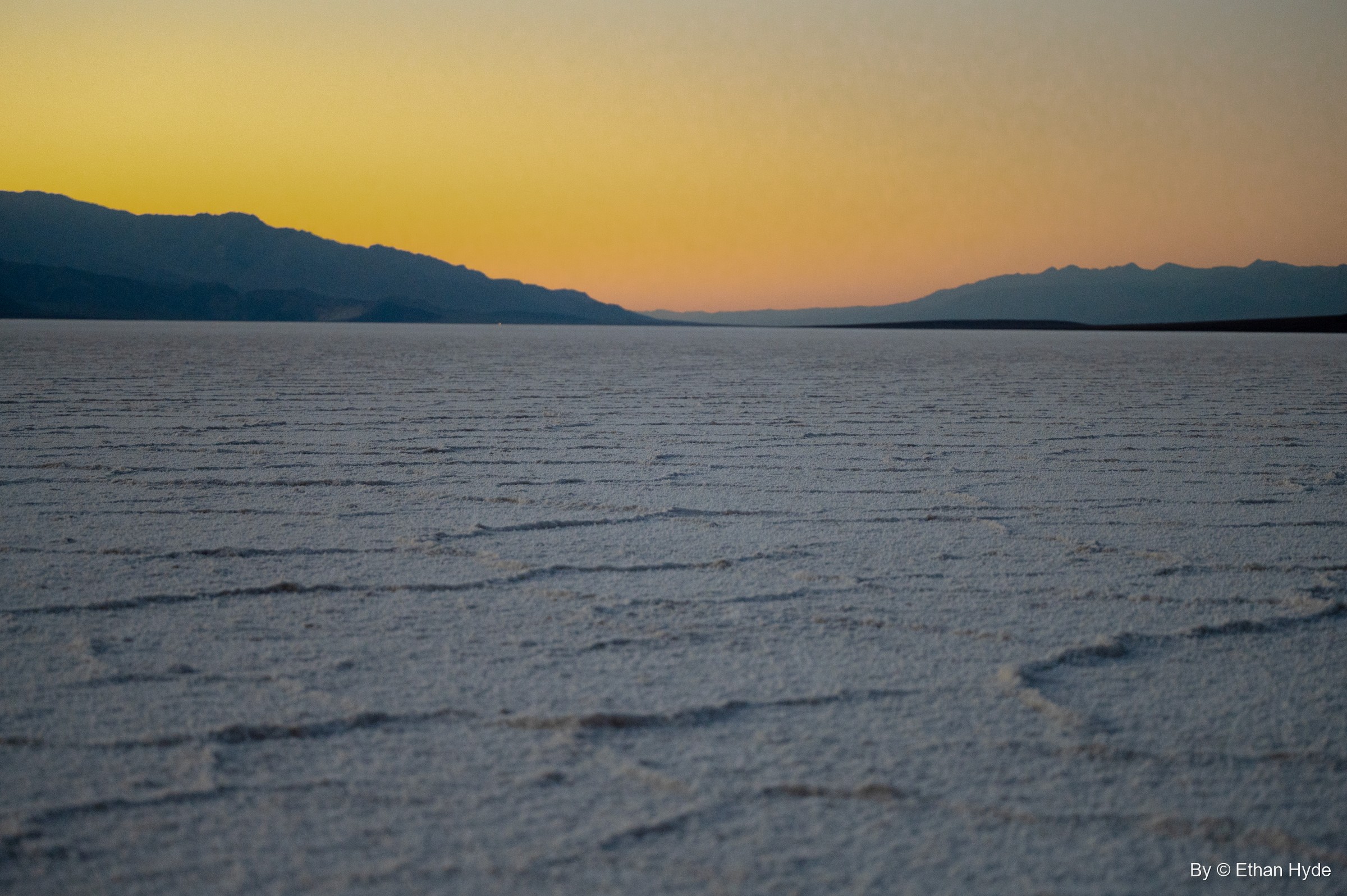 Ridged salt polygons stretch across Badwater Basin under an orange afterglow and a dark mountain silhouette.