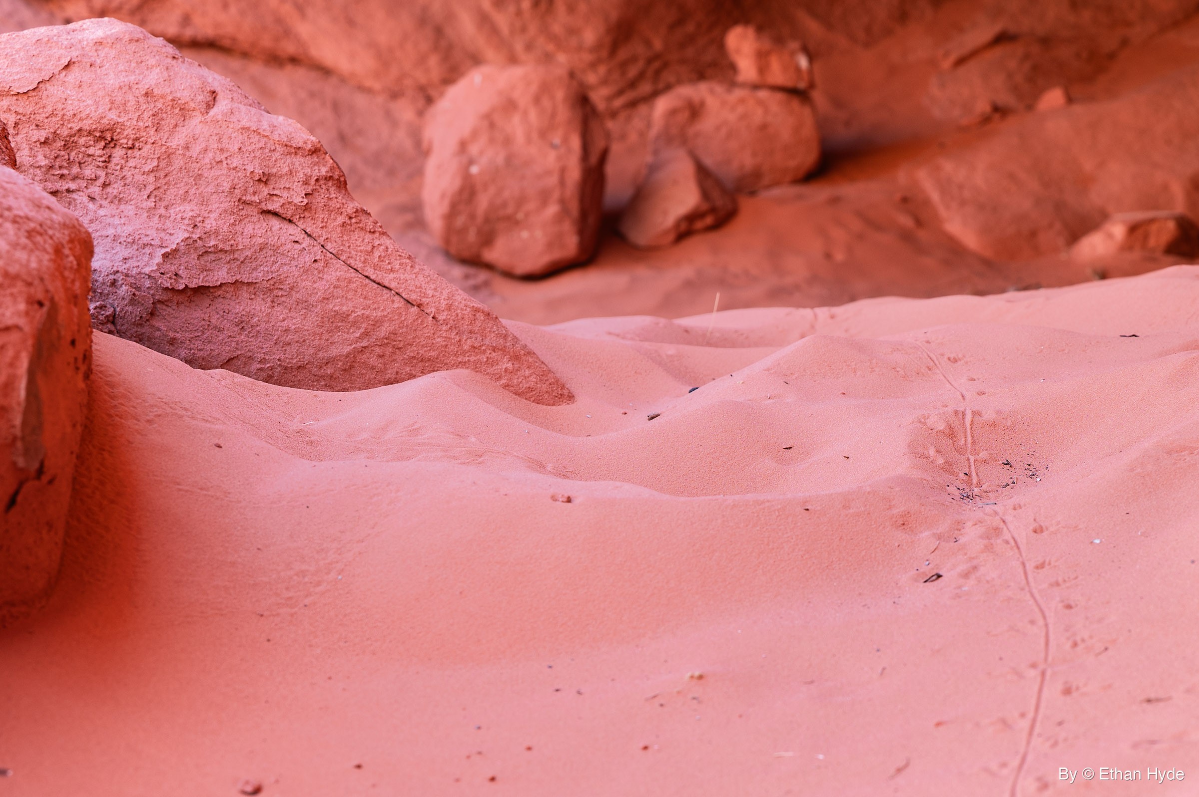 Mounded red sand spreads between sandstone boulders, with a thin line of small tracks and a tail drag along the right side.