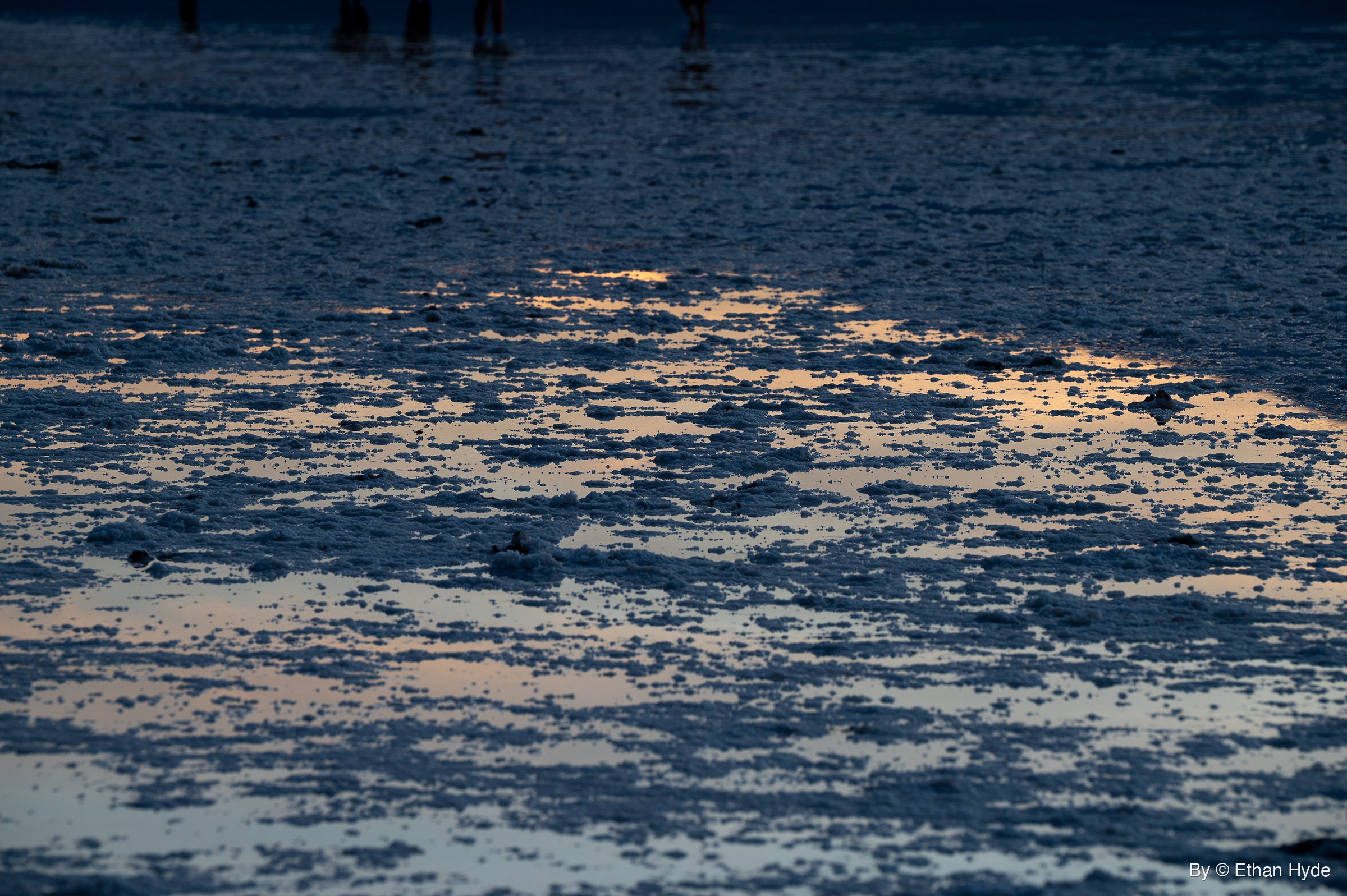 Shallow water pools between clumps of salt crust, reflecting an orange and pale blue sky, with the silhouetted legs of a few people at the top edge.