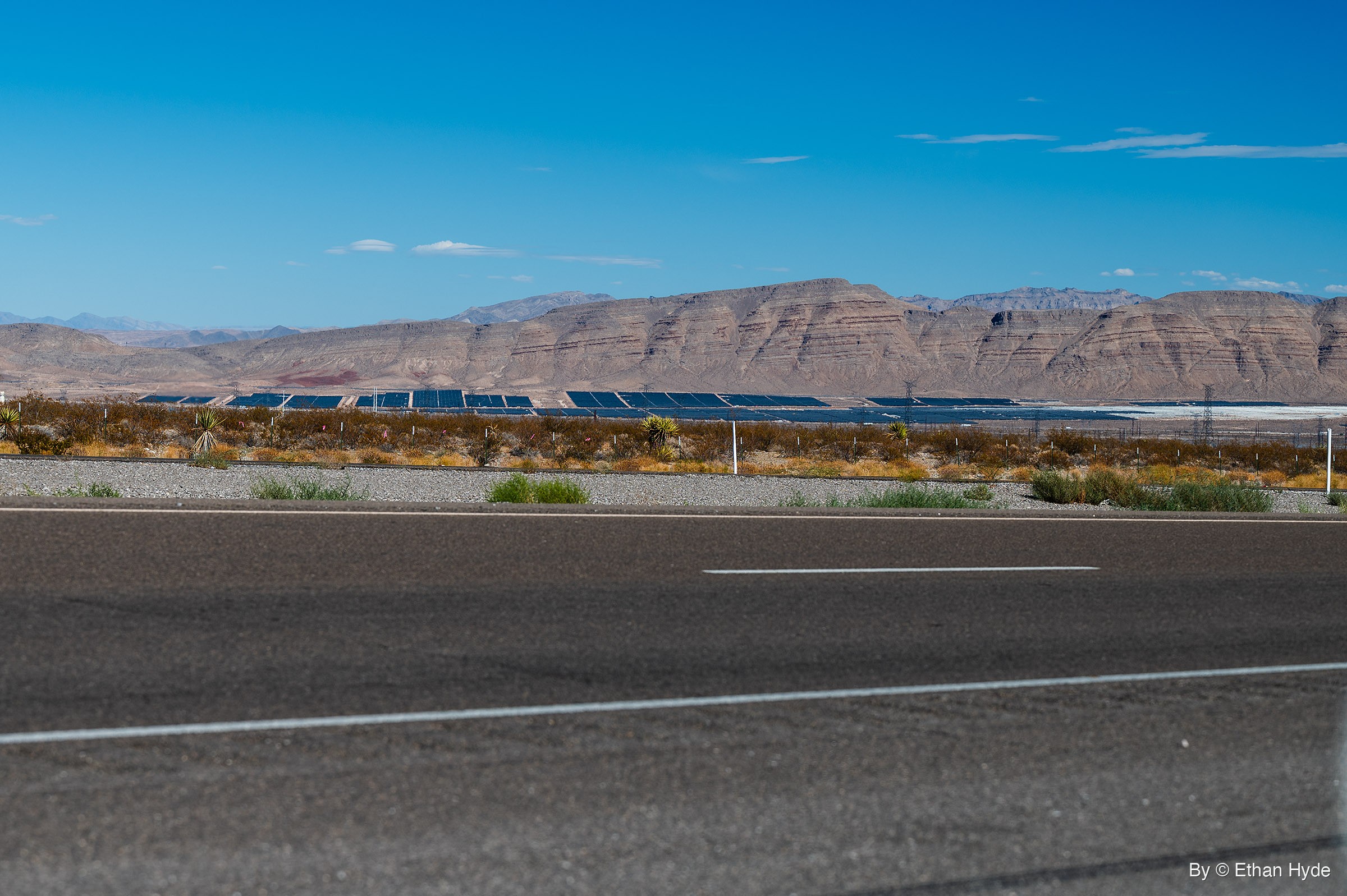 Rows of dark solar panels stretch across desert flats below layered tan ridges, seen past yucca scrub and a paved highway.