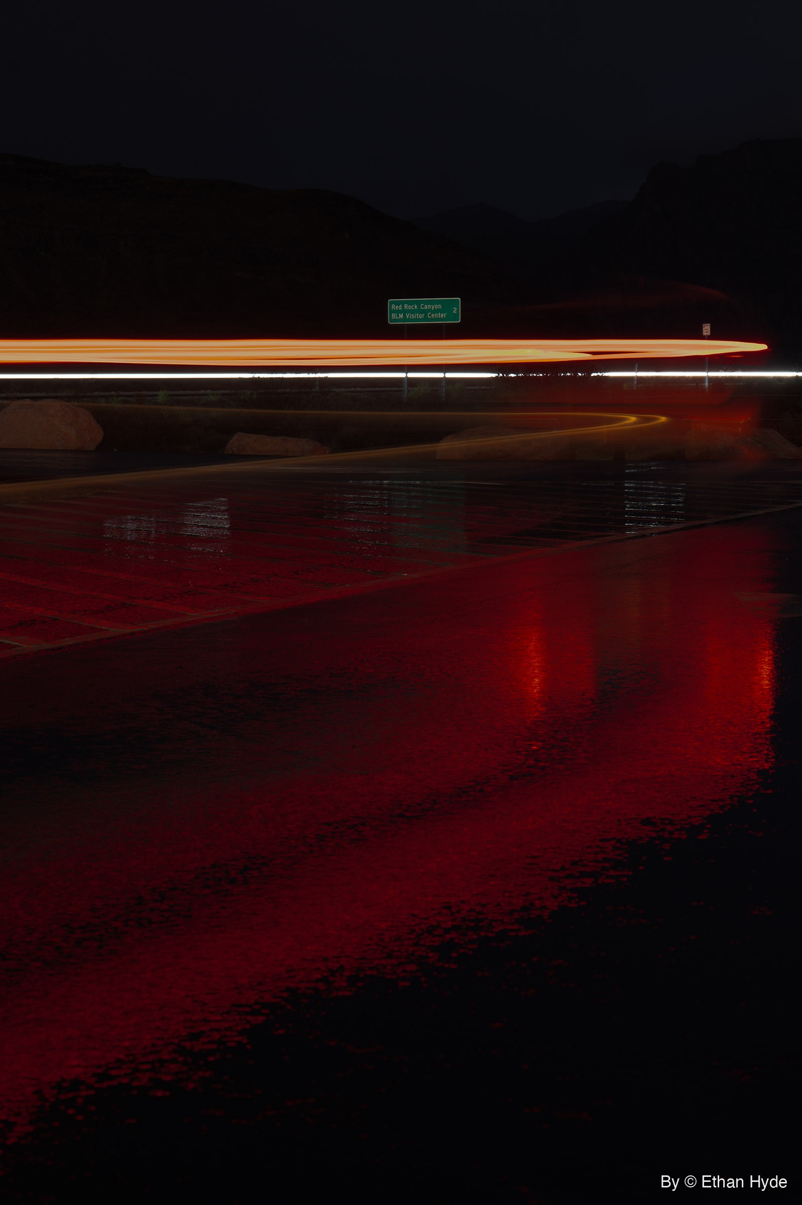 An orange taillight streak and a thin white light trail cross the frame at night above a rain-wet parking area glowing red, with dark mountains behind.