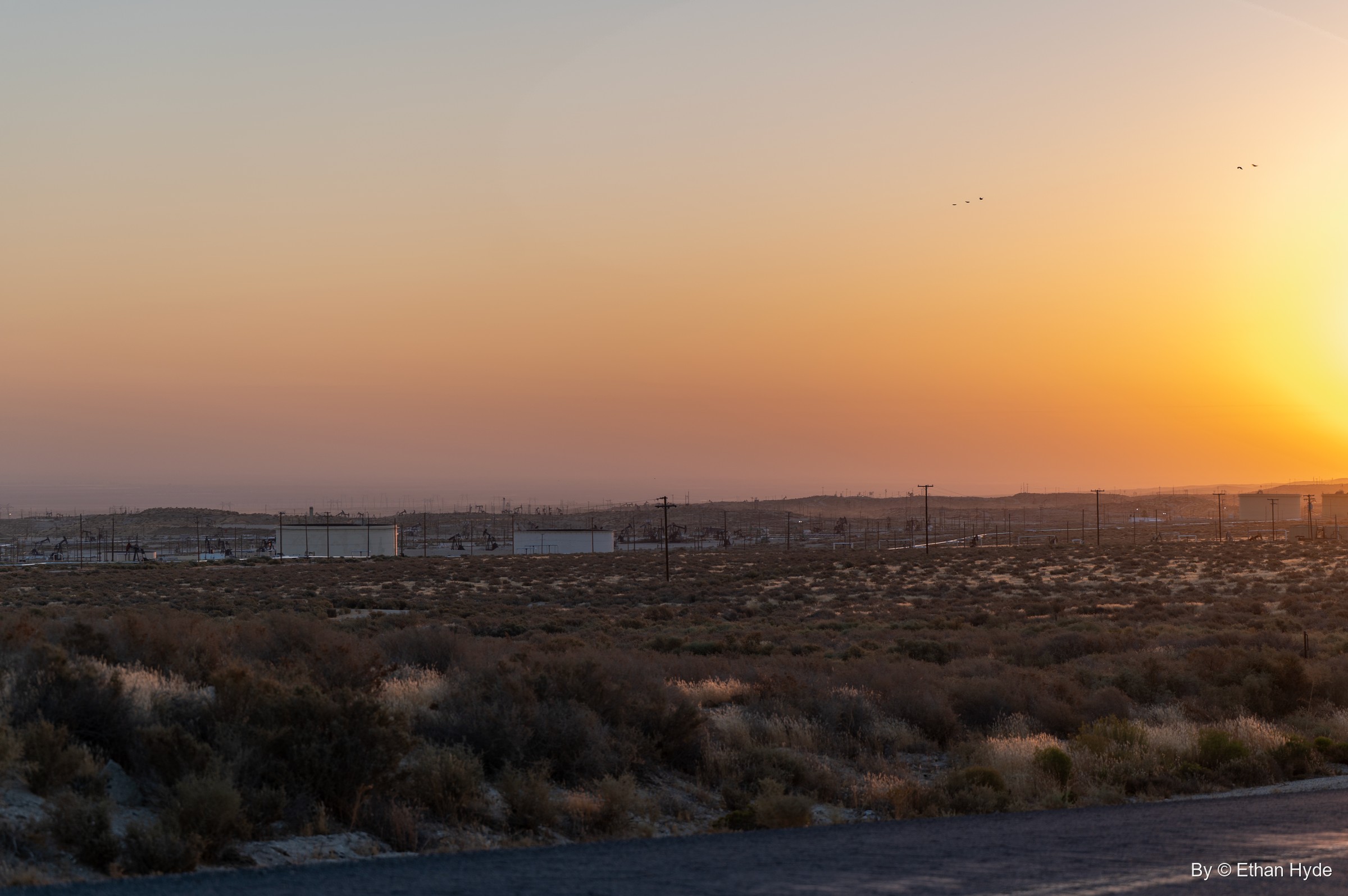 The sun breaks the horizon in an orange sky over desert scrub, with the oil field's tanks and power poles strung along the distance.