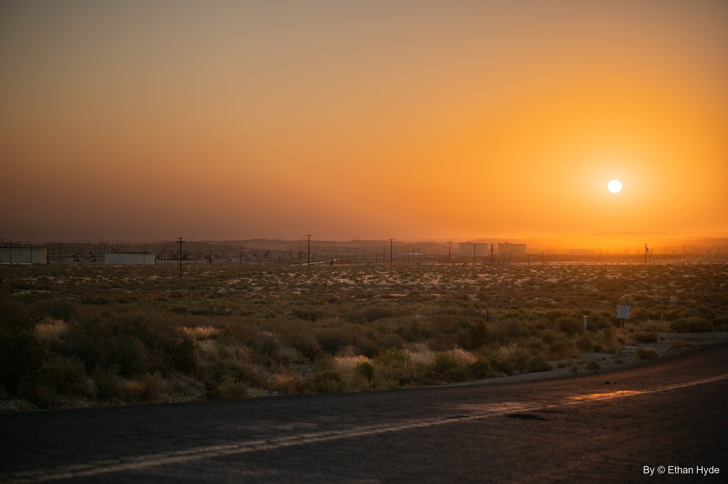 An orange sun rises in haze over desert scrub and the distant McKittrick oil field, a road curving through the foreground.