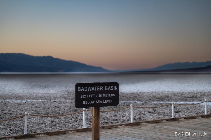 A brown sign reading "Badwater Basin, 282 feet / 86 meters below sea level" beside the boardwalk rail at dusk, the salt flat and mountains behind.