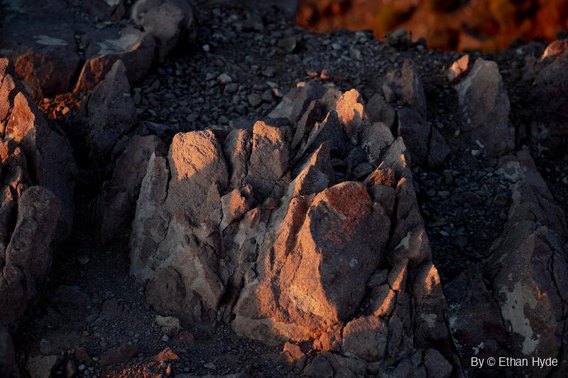 A rock outcrop broken into jagged upright blocks catches orange sunset light on a dark gravel slope, with a blurred orange hillside in the top corner.