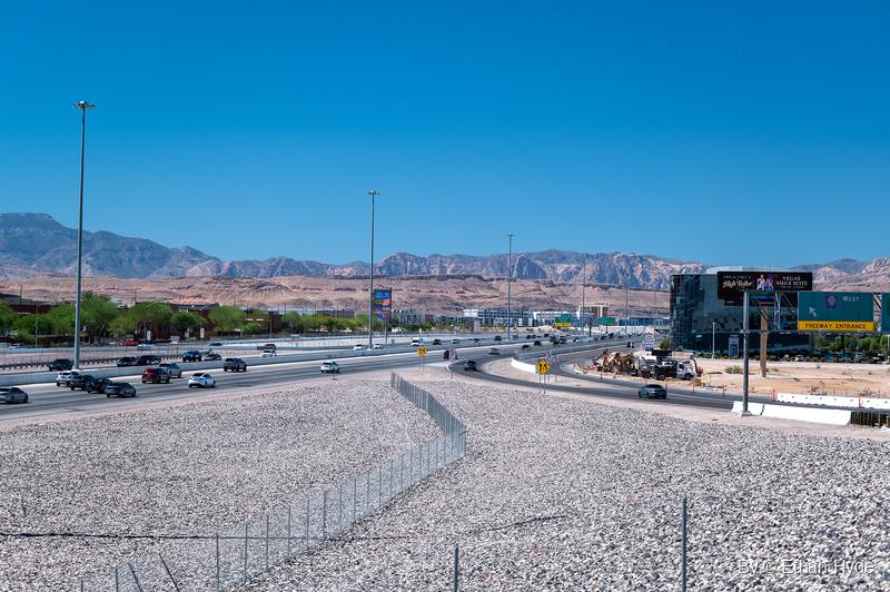 Wide view of a busy freeway and curving on-ramp beside a construction site, with a gravel slope and fence in front and rugged mountains beyond.