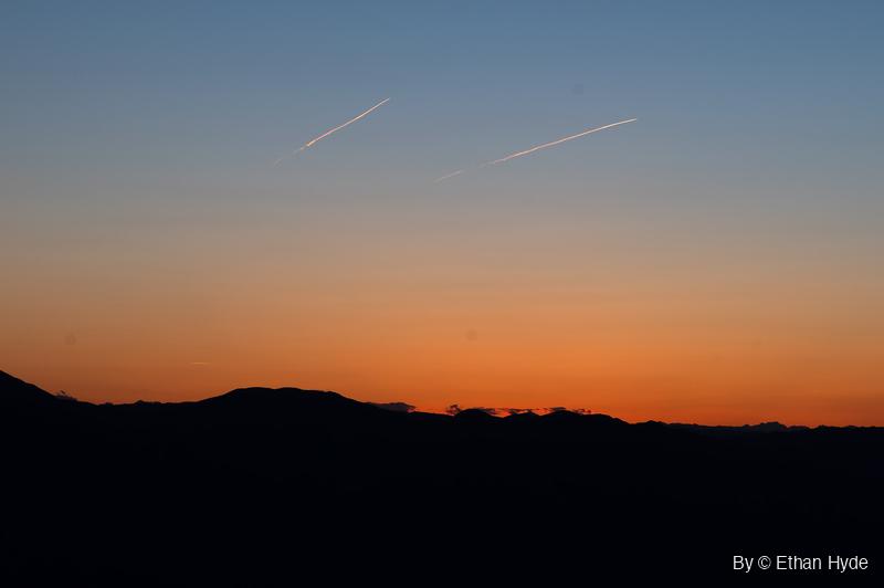 Two aircraft contrails cross a blue-to-orange twilight sky above a black silhouetted mountain ridge.