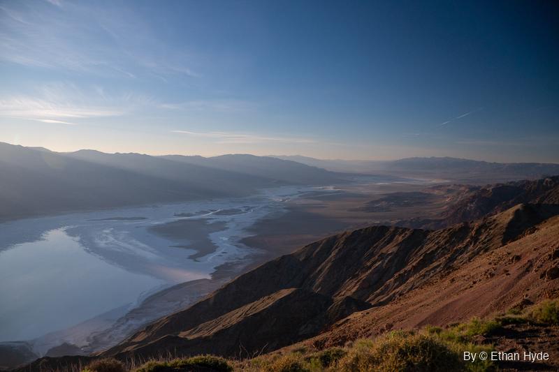 Looking north from a high brushy ridge down Death Valley, with shallow water spread across the salt flat and hazy ranges beyond.