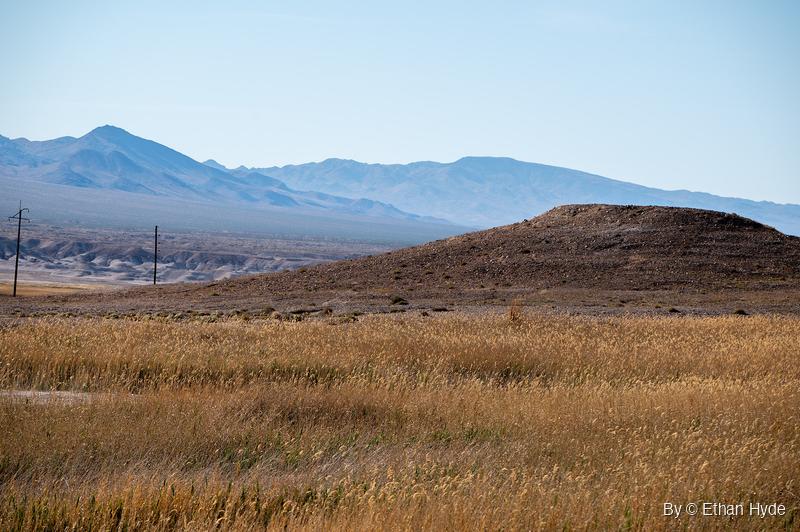 A field of dry golden grass in front of a brown dirt mound, with pale eroded badlands, two power poles and hazy blue mountains behind.