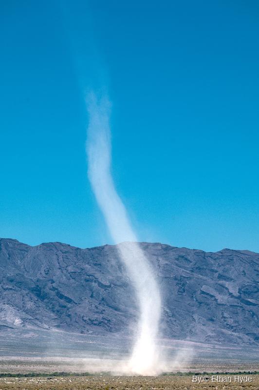 A tall pale dust devil rises from a desert floor, bending as it climbs past a gray mountain ridge into clear blue sky.