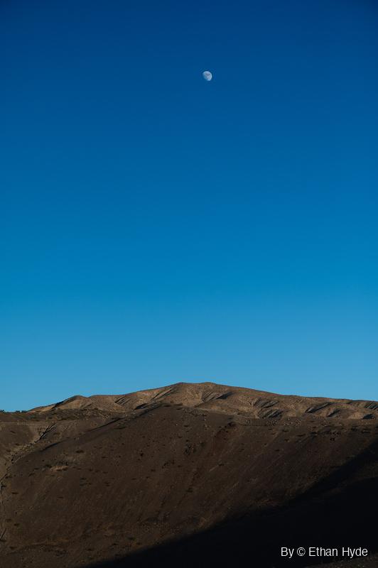 A small moon hangs high in a deep blue sky above a bare desert ridge whose near slope lies in shadow.