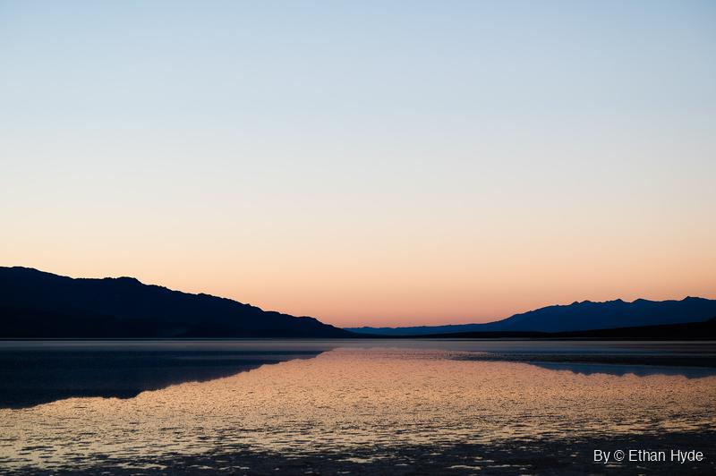 Shallow floodwater over the salt flat reflects an orange and blue sunset sky between two dark mountain ridges.