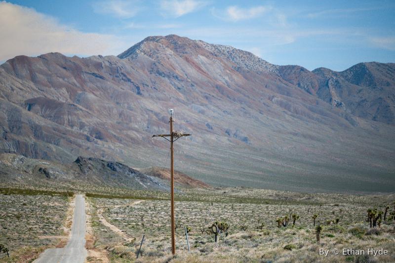 A dirt track and a lone power pole crossing a Joshua-tree valley toward mountains.