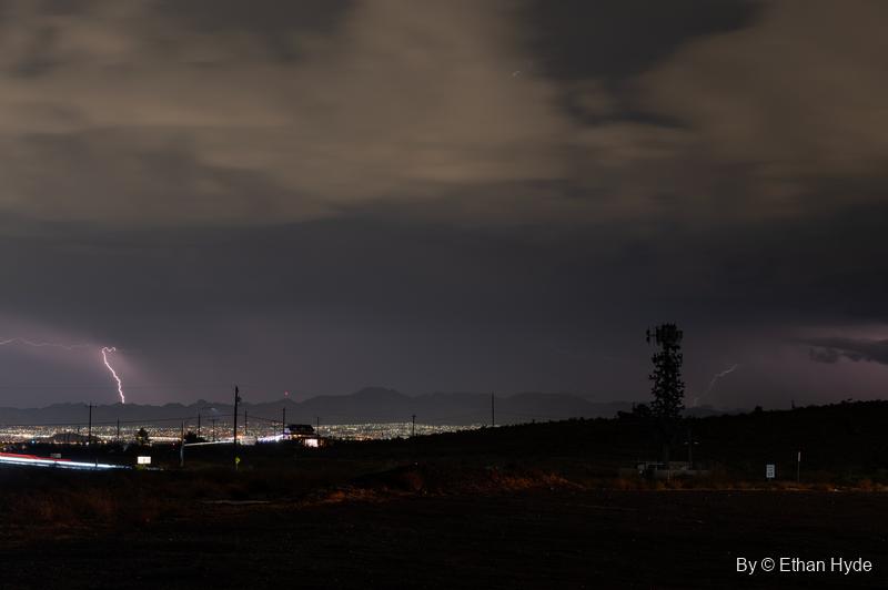 A lightning bolt strikes beyond a spread of city lights at night, with a cell tower silhouetted on the dark ridge.