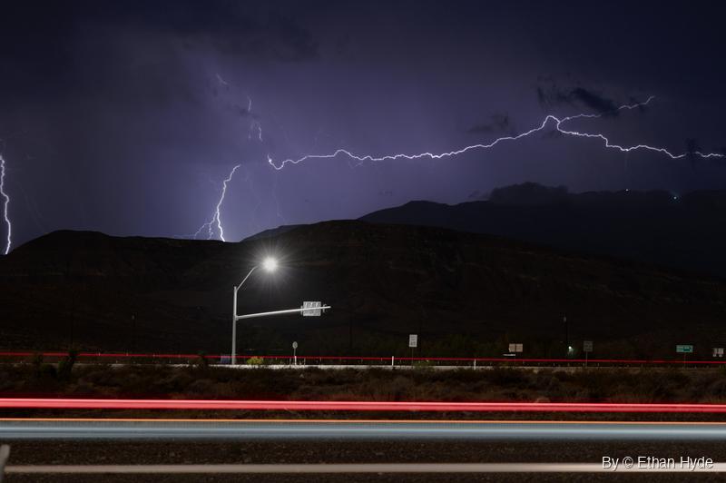 Lightning branches across a purple night sky above dark mountains, with a lit streetlight and red and white car light trails along a road below.