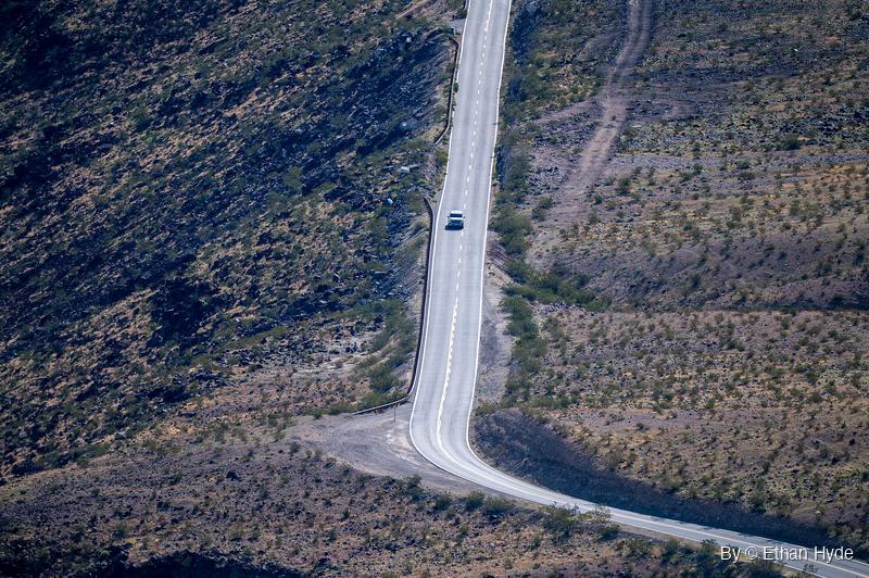 A single white car travels a two-lane road running straight up a scrubby desert slope, with a guardrail on the left and a curve at the bottom.