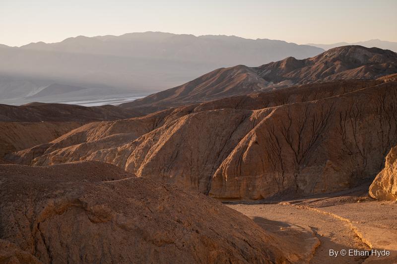 Low sun lights brown, eroded hills off Artists Drive, with the valley floor and a far mountain range in haze beyond.