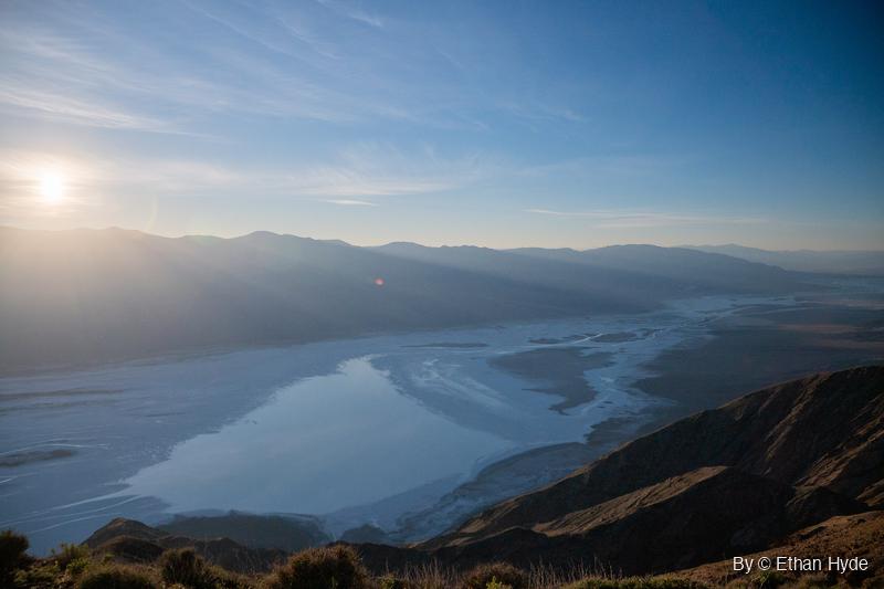 The sun hangs low over a hazy mountain range above Death Valley's salt flat, which is covered in shallow water, seen from a brushy ridge.