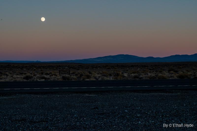 A near-full moon rises into a pink and blue dusk sky above a low mountain range, sagebrush flats and a paved highway with a gravel shoulder.