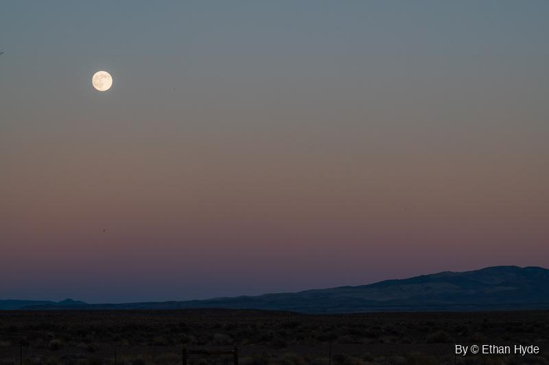 A nearly full moon high in a blue-to-pink twilight sky above a dark mountain ridge, a brushy flat and a fence line.