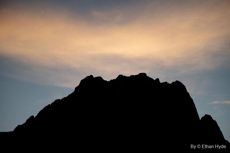 A jagged mountain peak in black silhouette under a warm, cloudy sunset sky.